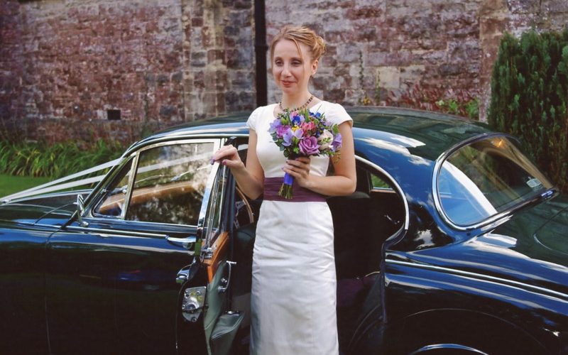 Bride next to wedding car, wearing short wedding dress with purple sash